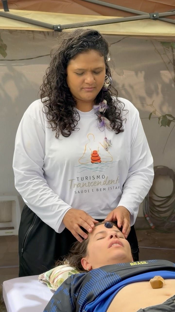 Terapeuta com camiseta do Turismo Transcendental conduzindo uma massagem com pedras terapêuticas na testa de uma pessoa